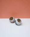 Two ceramic pots on a white surface with a peach-colored background. Liz Vidal Ceramics | Shop Fodder