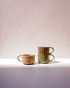 Three handmade ceramic mugs with rustic glazes in earthy tones of brown, green, and orange, arranged on a white surface against a soft beige background. Carved Mug | Scree Ceramics | Fodder