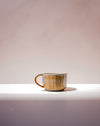 A handmade ceramic coffee mug with a rustic, textured glaze in earthy brown, beige, and gold tones, sitting on a white ledge against a soft, warm beige background. Carved Mug | Scree Ceramics | Fodder
