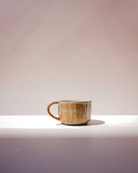 A handmade ceramic coffee mug with a rustic, textured glaze in earthy brown, beige, and gold tones, sitting on a white ledge against a soft, warm beige background. Carved Mug | Scree Ceramics | Fodder