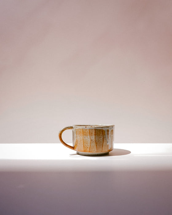 A handmade ceramic coffee mug with a rustic, textured glaze in earthy brown, beige, and gold tones, sitting on a white ledge against a soft, warm beige background. Carved Mug | Scree Ceramics | Fodder