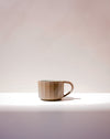 A small ceramic mug with a faceted, geometric design in a warm beige and brown glaze, sitting on a white surface against a soft beige background. Carved Mug | Scree Ceramics | Fodder