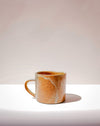 A small handmade ceramic mug with a warm orange-brown glaze and white marbling, sitting on a white surface against a light beige background. Everyday Mug | Scree Ceramics | Fodder