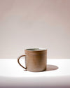 A handmade ceramic mug with a textured, earthy brown exterior and a speckled light green interior, sitting on a white surface against a warm beige background. Everyday Mug | Scree Ceramics | Fodder