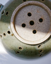 Close-up of the underside of a ceramic vessel, showing a round beige base with five drainage holes and a green glazed exterior with multiple side ventilation holes. handmade ceramic colander by Scree Ceramics | Fodder