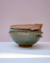 Three stacked handmade ceramic bowls with a speckled glaze, featuring a top bowl with a raw, unglazed rim, displayed on a white surface against a soft pink background.