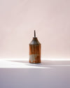 A small ceramic bottle with a conical shape, featuring a textured glaze in orange, brown, and cream, with a dark spout, set against a soft pink background with a white surface below. Oil Bottle | Scree Ceramics | Fodder