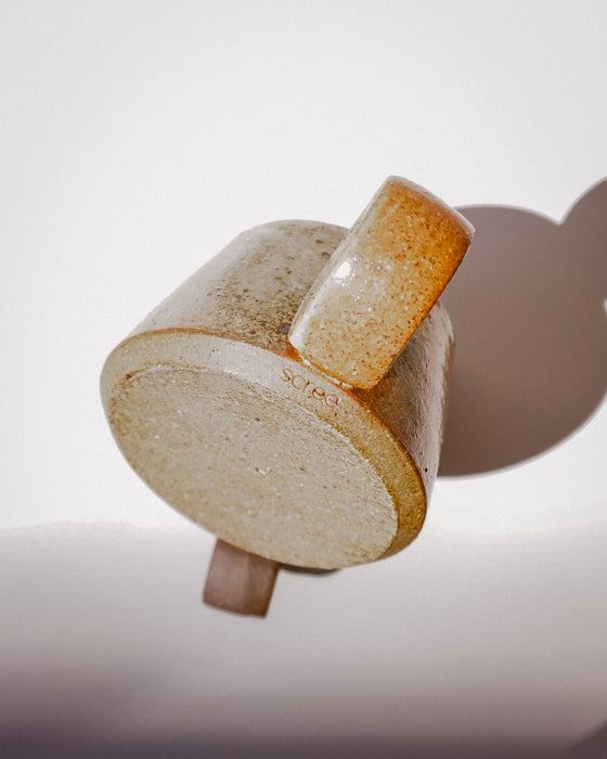 A close-up studio shot of a ceramic mug showing its textured beige and brown glaze, side handle, and base detail against a white background. Short Loop Mug | Scree Ceramics | Fodder