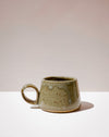 A handmade ceramic mug with a rounded body, a small loop handle, and a textured, speckled glaze in earthy olive green and beige tones, set against a minimalist beige and white background. Short Loop Mug | Scree Ceramics | Fodder