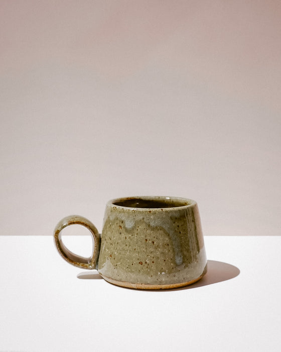 A handmade ceramic mug with a rounded body, a small loop handle, and a textured, speckled glaze in earthy olive green and beige tones, set against a minimalist beige and white background. Short Loop Mug | Scree Ceramics | Fodder