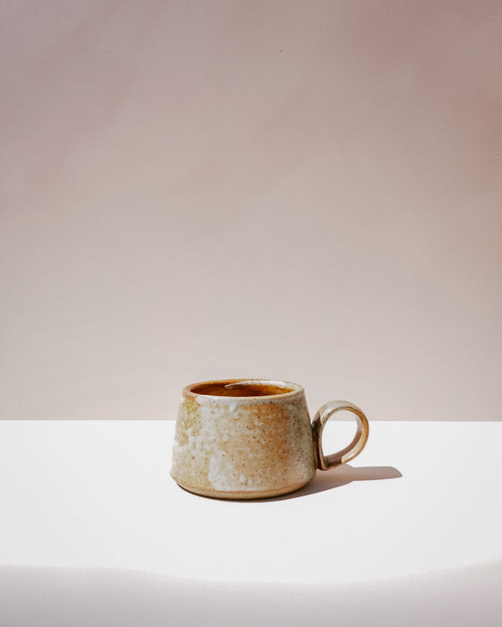 A small handmade ceramic mug with a textured, speckled beige and brown glaze and a C-shaped handle, sitting on a white surface against a warm beige background. Short Loop Mug | Scree Ceramics | Fodder