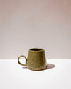 A small handmade ceramic mug with a tapered shape and a loop handle, featuring a textured olive green glaze with brown speckles, set against a minimalist beige and white background. Tall Loop Mug | Scree Ceramics | Fodder