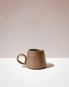 A small handmade ceramic mug with a tapered shape and a loop handle, featuring a textured earthy brown exterior and a dark green glazed interior, set against a minimalist beige and white background. Tall Loop Mug | Scree Ceramics | Fodder