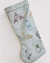 A light gray fabric Christmas stocking with embroidered holiday-themed illustrations including birds, a milk bottle, a swan, a glass, a chicken, and a ring, shown on a plain white background.12 Days of Christmas embroidered linen Christmas stocking by See & Quin - carol motifs - Fodder