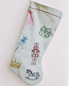 A light blue fabric Christmas stocking decorated with colorful embroidered toy-themed motifs including a nutcracker, drum, ballet shoes, wand, crown, and rocking horse, shown against a plain off-white background. Nutcracker embroidered linen Christmas stocking by See & Quin - ballet motifs - Fodder