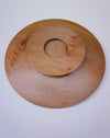 A round wooden serving board with a textured grain, featuring a concentric circular cutout in the center, photographed from directly above on a light gray background.