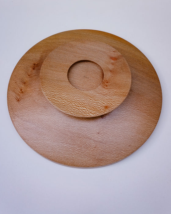A round wooden serving board with a textured grain, featuring a concentric circular cutout in the center, photographed from directly above on a light gray background.
