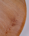 A close-up of a round, textured surface in warm tan and brown tones with a fine ribbed pattern and a raised circular edge, shown against a light neutral background. London Plane wooden cake stand by Selwyn House - lacewood grain - Fodder