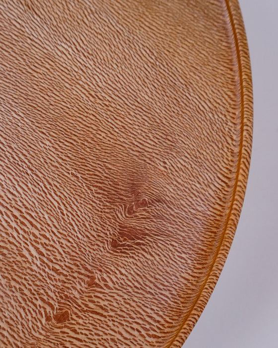 A close-up of a round, textured surface in warm tan and brown tones with a fine ribbed pattern and a raised circular edge, shown against a light neutral background. London Plane wooden cake stand by Selwyn House - lacewood grain - Fodder