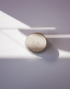 Round beige object with a brand logo on a light purple background. Sophie Alda | Handmade Ceramics | Shop Fodder