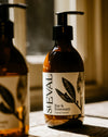 An amber glass pump bottle of St Eval Bay & Rosemary hand lotion on a wooden surface, with a softly blurred second bottle in the background. Bay & Rosemary hand lotion by St Eval - refillable glass bottle - Fodder
