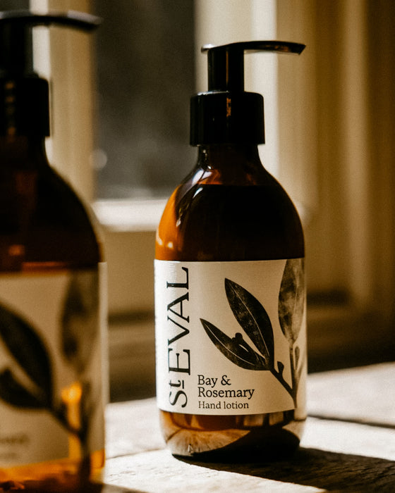 An amber glass pump bottle of St Eval Bay & Rosemary hand lotion on a wooden surface, with a softly blurred second bottle in the background. Bay & Rosemary hand lotion by St Eval - refillable glass bottle - Fodder
