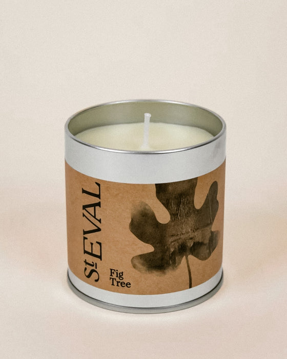 A StEVAL Fig Tree scented candle in a silver metal tin with a brown kraft paper label featuring a dark leaf illustration, set against a solid light beige background.
