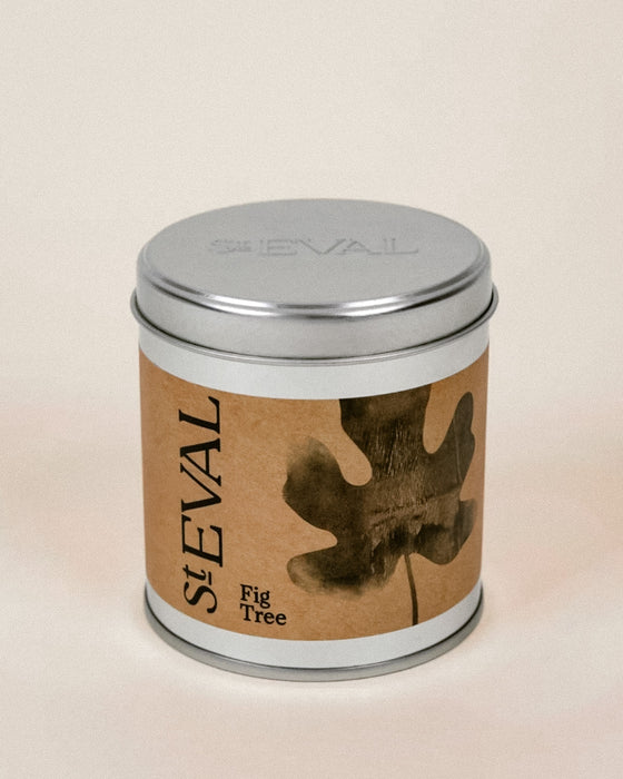 A cylindrical metal tin candle with a silver lid and a brown kraft paper label featuring a dark leaf graphic and the text 'st EVAL Fig Tree'. Fig Tree scented tin candle by St Eval - illustrated tin - Fodder