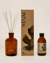 Seval Fig Tree scented oil diffuser set including an amber glass reed diffuser bottle with reeds, a tall brown kraft paper box, and a smaller amber glass bottle with a black cap, all on a warm beige background. Fig Tree reed diffuser by St Eval - fresh green home fragrance - Fodder
