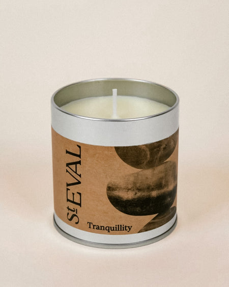 A single-wick candle in a cylindrical silver metal tin with a kraft paper label reading 'StEVAL Tranquillity' and a dark abstract moon graphic, set against a warm beige background.