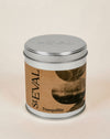 A cylindrical metal tin candle with a silver lid and a brown paper label reading 'StEVAL Tranquillity' with abstract dark circular artwork, photographed on a plain light beige background.