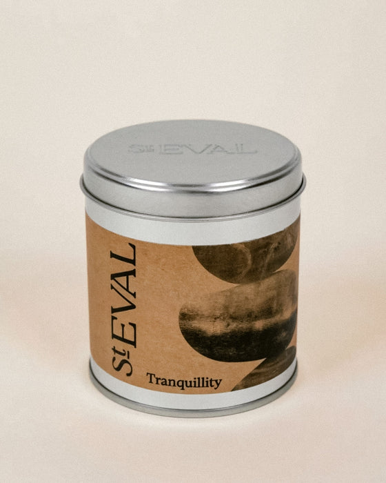 A cylindrical metal tin candle with a silver lid and a brown paper label reading 'StEVAL Tranquillity' with abstract dark circular artwork, photographed on a plain light beige background.