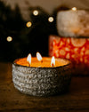 A lit three-wick candle in a textured gray ceramic holder sits on a wooden surface, with warm blurred lights and additional candles in the background.