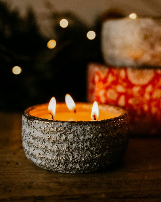 A lit three-wick candle in a textured gray ceramic holder sits on a wooden surface, with warm blurred lights and additional candles in the background.