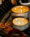 Two St Eval lit candles in textured grey ceramic bowls sit on a dark tray with dried orange slices nearby. The scene is warmly lit, with the candles as the main product and no people or animals present.