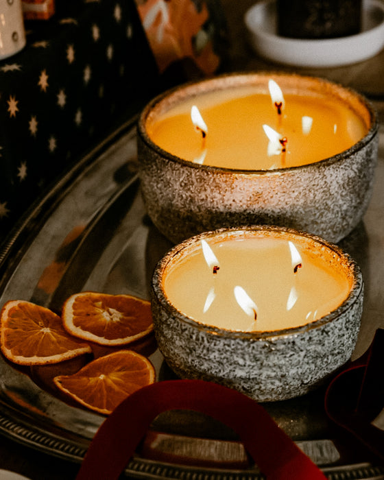 Two St Eval lit candles in textured grey ceramic bowls sit on a dark tray with dried orange slices nearby. The scene is warmly lit, with the candles as the main product and no people or animals present.