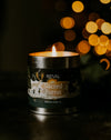 A lit candle in a matte black metal tin labeled 'St EVAL Sacred Forest' sits on a dark surface with warm golden bokeh lights blurred in the background.