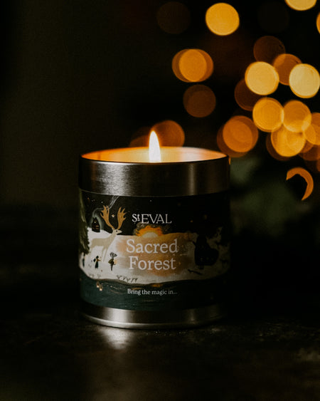 A lit candle in a matte black metal tin labeled 'St EVAL Sacred Forest' sits on a dark surface with warm golden bokeh lights blurred in the background.