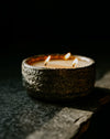 A three-wick candle in a rustic, textured dark brown ceramic holder, lit with three small flames against a dark, moody background.