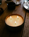 A three-wick candle in a dark, rustic ceramic bowl, lit with three visible flames on a wooden table.
