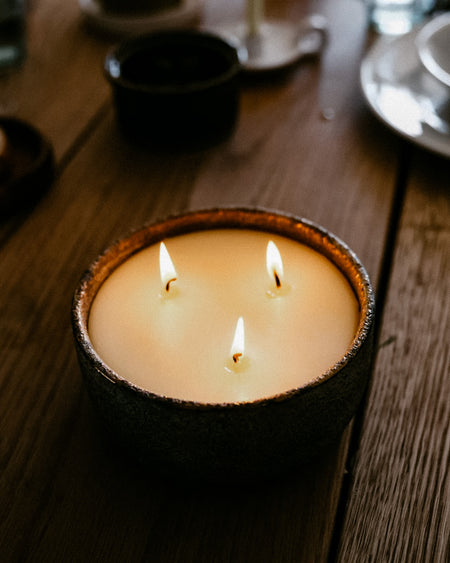 A three-wick candle in a dark, rustic ceramic bowl, lit with three visible flames on a wooden table.