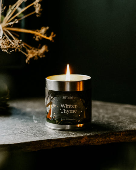 A lit SEVAL Winter Thyme candle in a silver metal tin with a dark label, set on a dark stone surface with dried botanicals in the background.