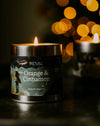 A lit metal tin candle labeled 'stEVAL Orange & Cinnamon' sits on a dark surface with warm blurred bokeh lights in the background.
