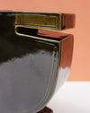 Close-up of a glossy ceramic bowl with a modern geometric design, featuring a dark charcoal-black body and a metallic olive-gold rim, set against a warm peach background. Studio ERS | Soup Vase | Fodder