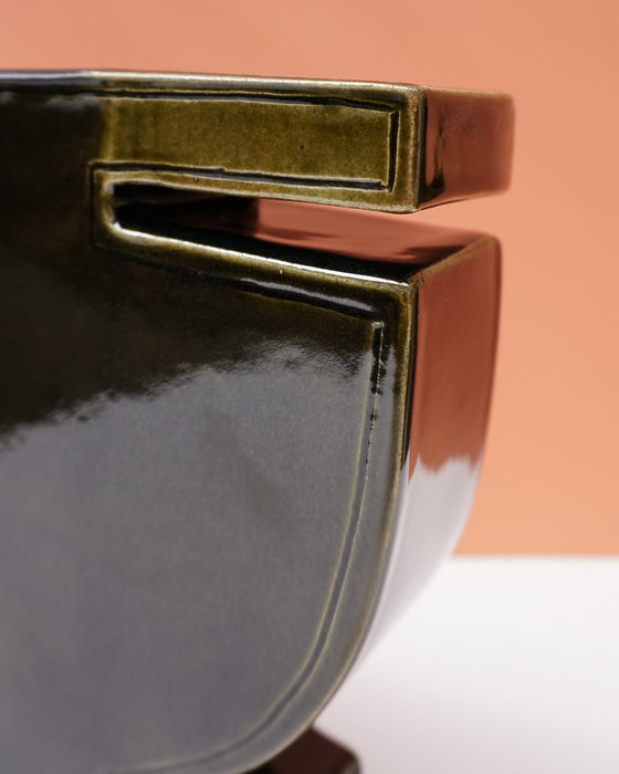 Close-up of a glossy ceramic bowl with a modern geometric design, featuring a dark charcoal-black body and a metallic olive-gold rim, set against a warm peach background. Studio ERS | Soup Vase | Fodder