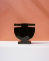 A glossy black ceramic vase with a wide, rounded body and a small rectangular handle, displayed on a white surface against a warm peach-colored background. Studio ERS | Soup Vase | Fodder