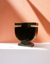 A glossy black ceramic urn-shaped vase with a wide top rim, displayed on a white surface against a terracotta-colored background with sharp diagonal shadows. Studio ERS | Soup Vase | Fodder
