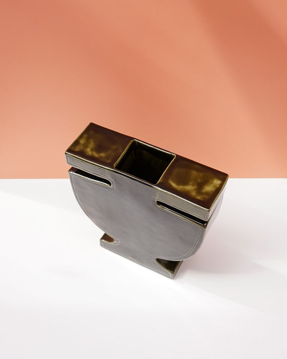 A modern sculptural metal container or candle holder with a rectangular top, a central square opening, and two side slots, shown on a white surface against a peach-colored background. Studio ERS | Soup Vase | Fodder