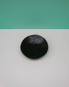 Black ceramic plate on a gray surface with a teal background. Diddy Bowl | Jode Pankhurst | Shop Fodder