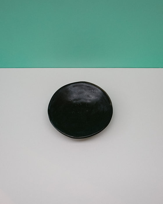 Black ceramic plate on a gray surface with a teal background. Diddy Bowl | Jode Pankhurst | Shop Fodder